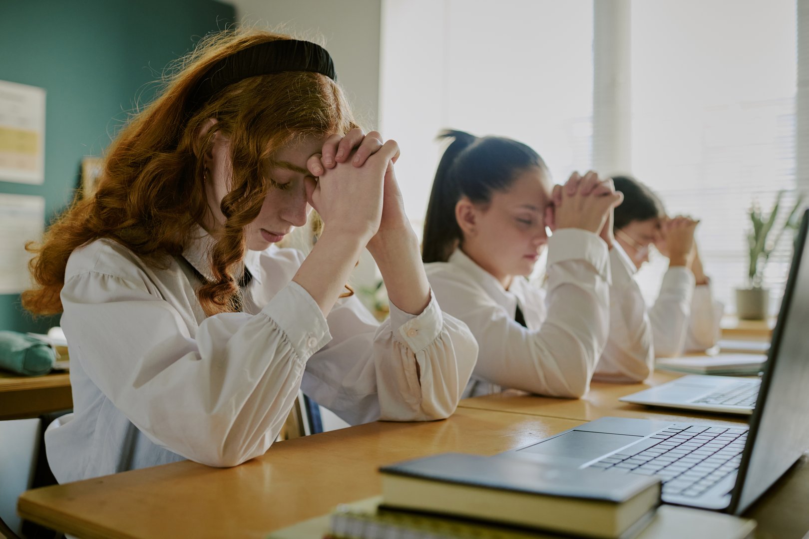 Group of students in school uniforms praying with closed eyes and folded hands while seated at desk in classroom environment