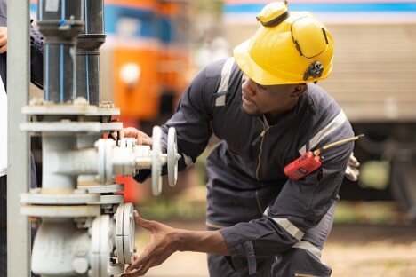 Engineer in a yellow hard hat adjusts valves on industrial pipes outdoors, focusing on equipment operation.