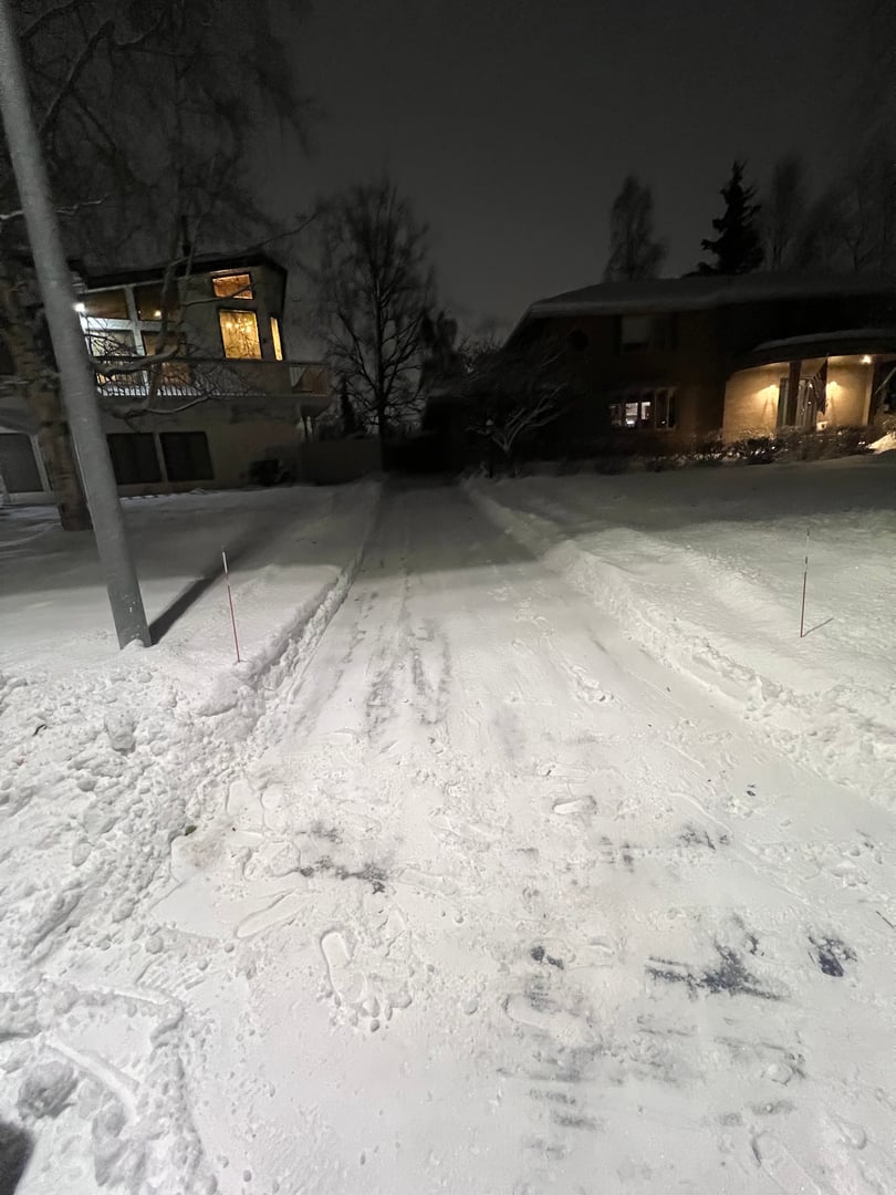 Residential driveway snow removal service showcasing cleared path in Anchorage, Alaska, with snow-covered surroundings and illuminated homes at night.