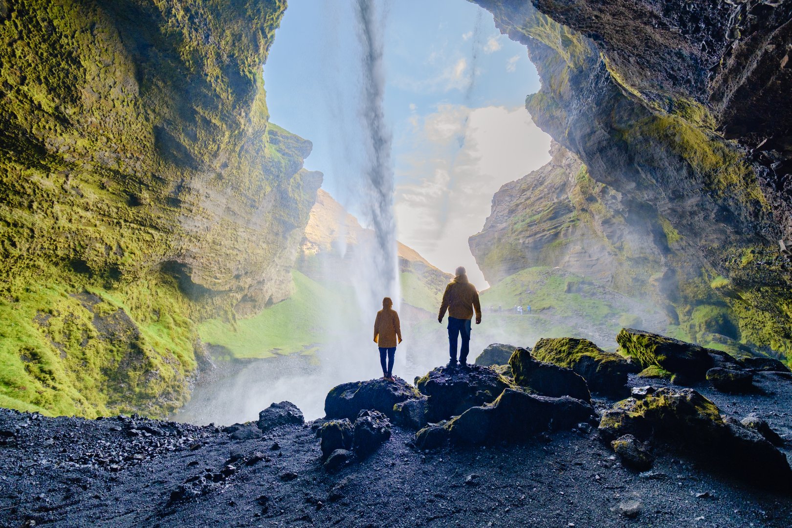 Two adventurers stand silhouetted against the breathtaking Kvernufoss waterfall, surrounded by dramatic cliffs and lush greenery.