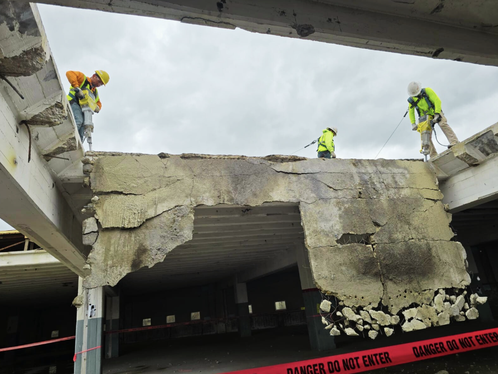 Construction workers demolishing a concrete structure with safety gear. Warning tape marks restricted area. Cloudy sky overhead.