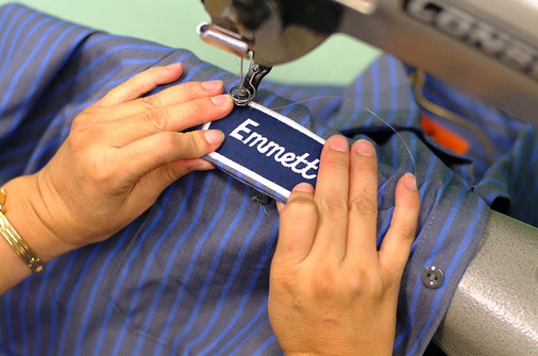 Person sewing a name patch labeled Emmett onto a blue striped shirt using a sewing machine.