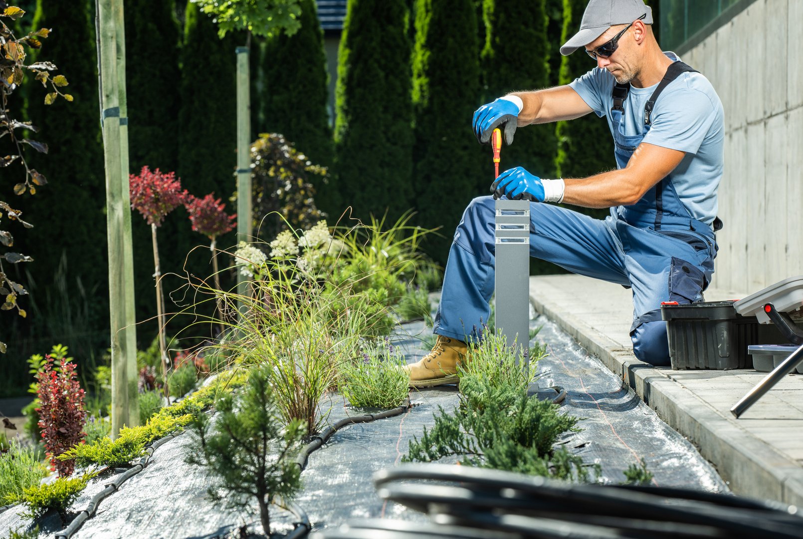 Caucasian Male Landscape Electrician with Opened Toolbox Next to Him Installing Backyard Garden Illumination System Screwing the Lamp with a Screwdriver. Thuja Wall in the Background.