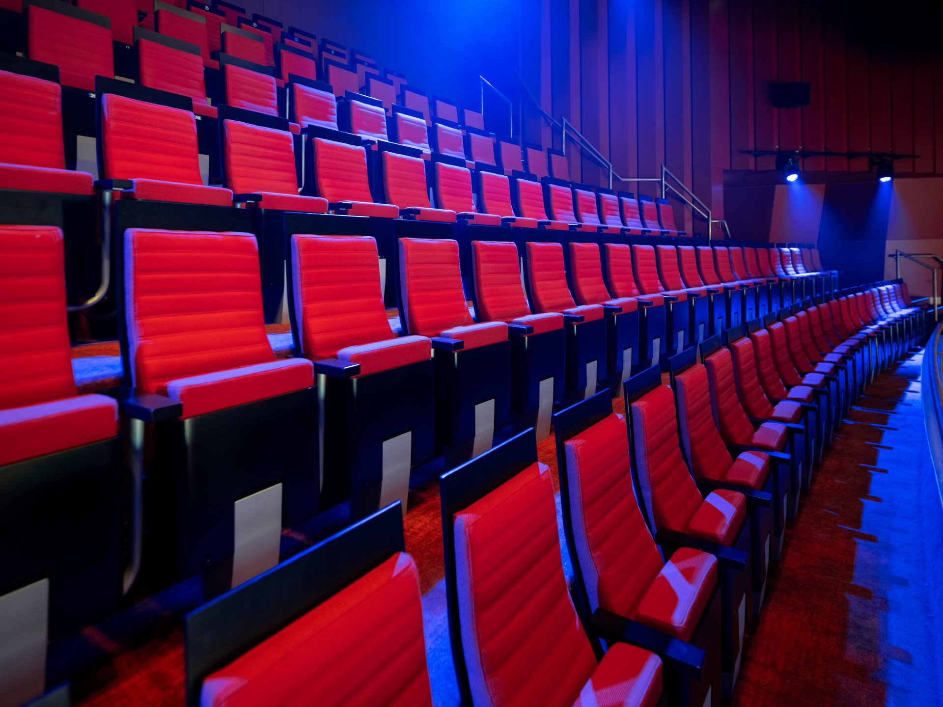 Rows of empty red theater seats with black frames line the auditorium under dramatic blue stage lighting