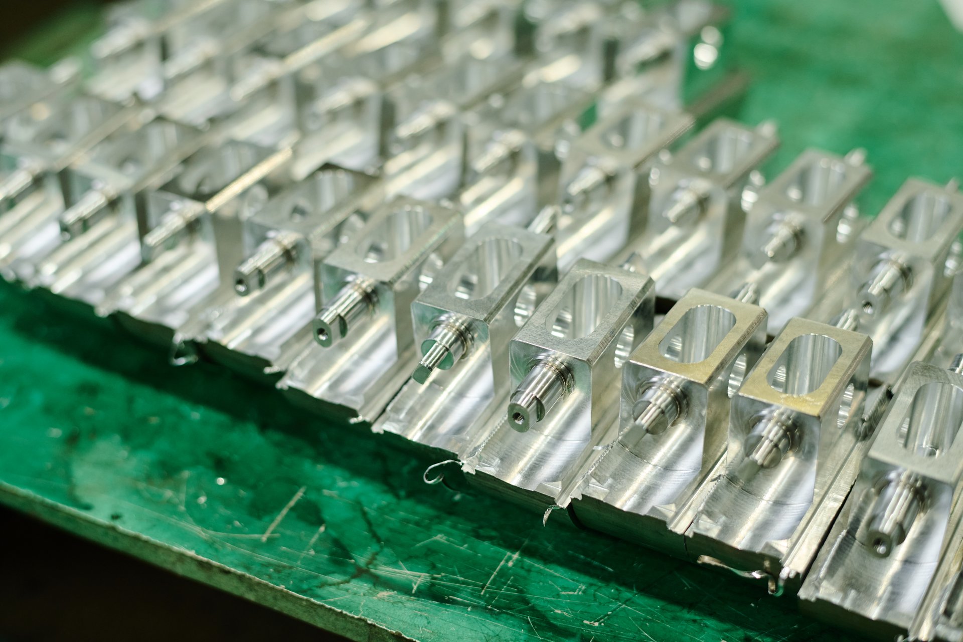Close-up of intricately designed industrial metal parts arranged in rows on green surface showcasing precision machining and manufacturing. Detailed view highlighting metallic sheen and uniformity