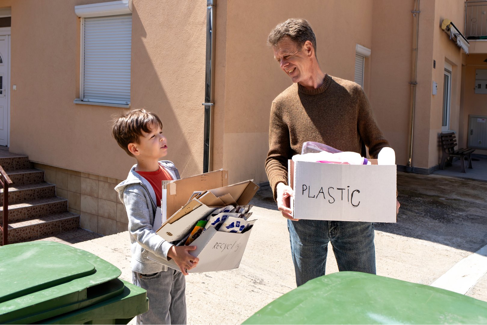 A father and son talk next to garbage bins while holding boxes with sorted waste. Concepts: children's involvement in household chores, eco-friendly family habits, sustainable lifestyle, waste sorting.