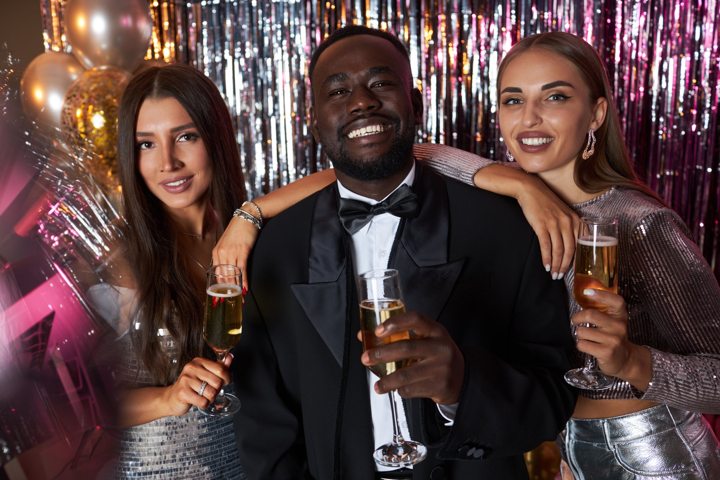 Three friends dressed elegantly, celebrating an event with drinks, sparkling decorations, and balloons, smiling warmly in front of the camera