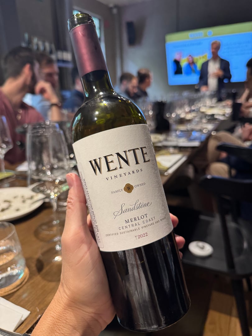 Hand holding a bottle of Wente Vineyards Sandstone Merlot 2022 at a dinner event with people and glasses in the background.