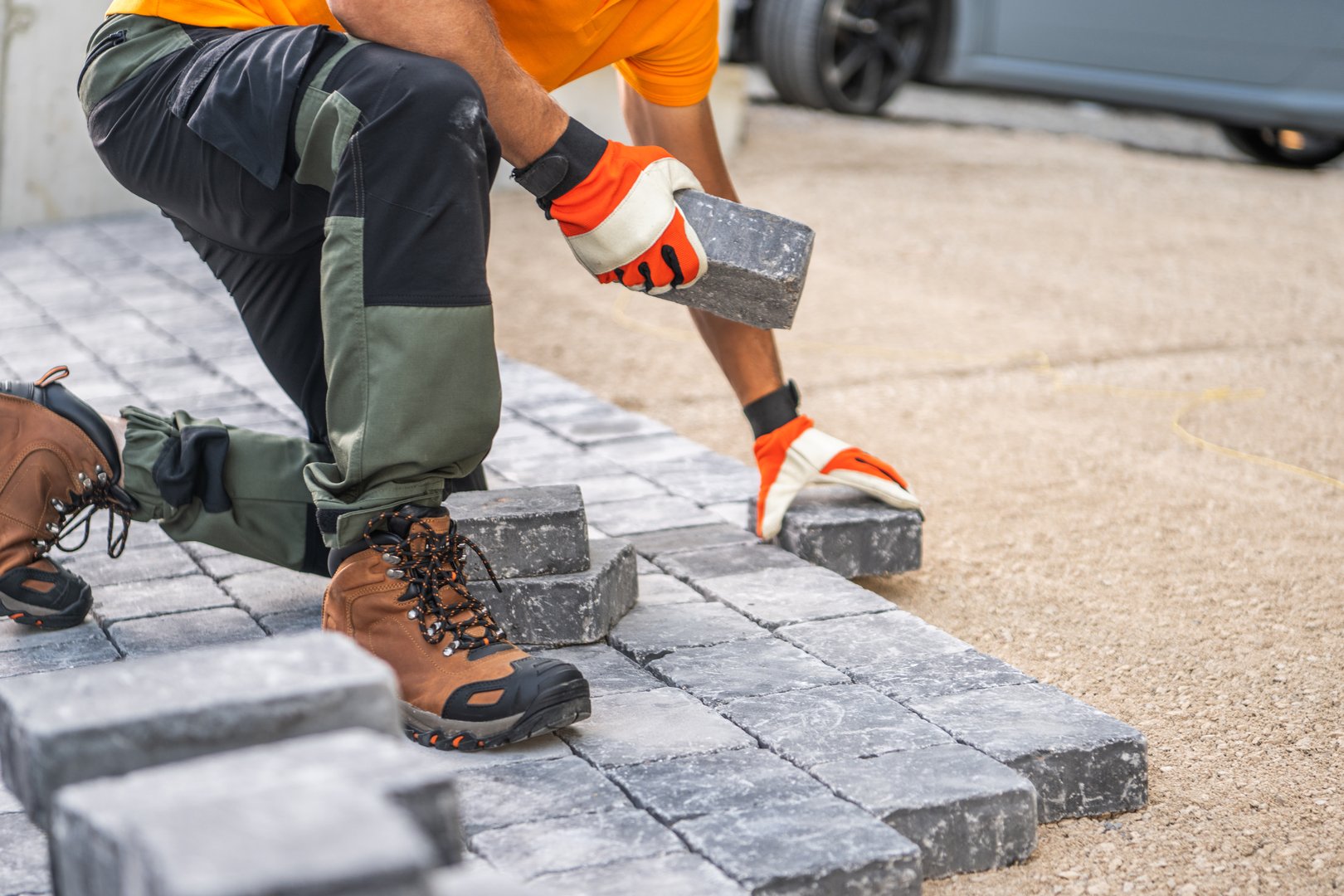 A construction worker is kneeling on the ground, carefully placing paving stones in a driveway. Wearing gloves and sturdy boots, they focus on arranging the stones for a neat, durable surface under clear skies.