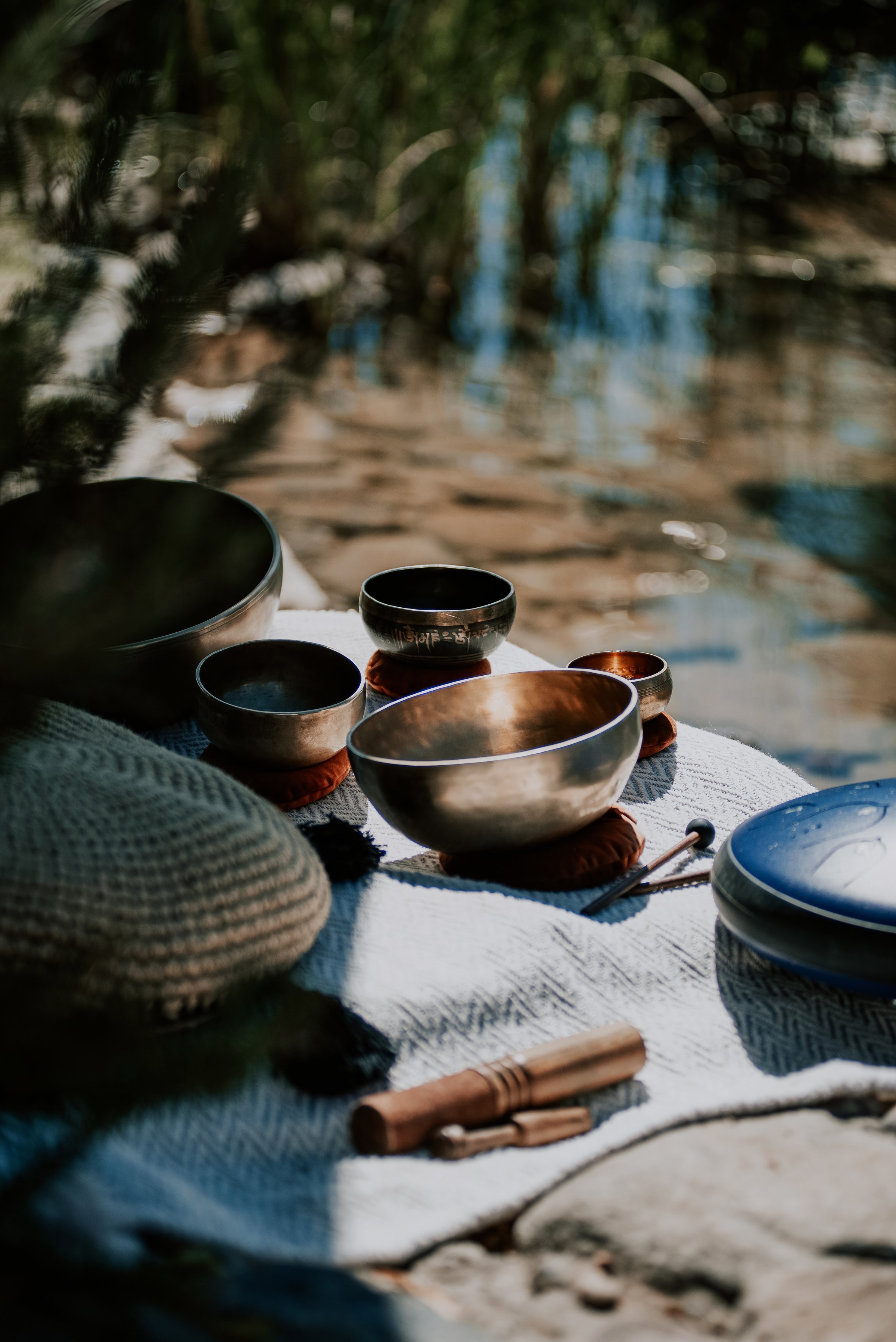 Several Tibetan singing bowls with mallets arranged on a blanket near a pond, surrounded by greenery.