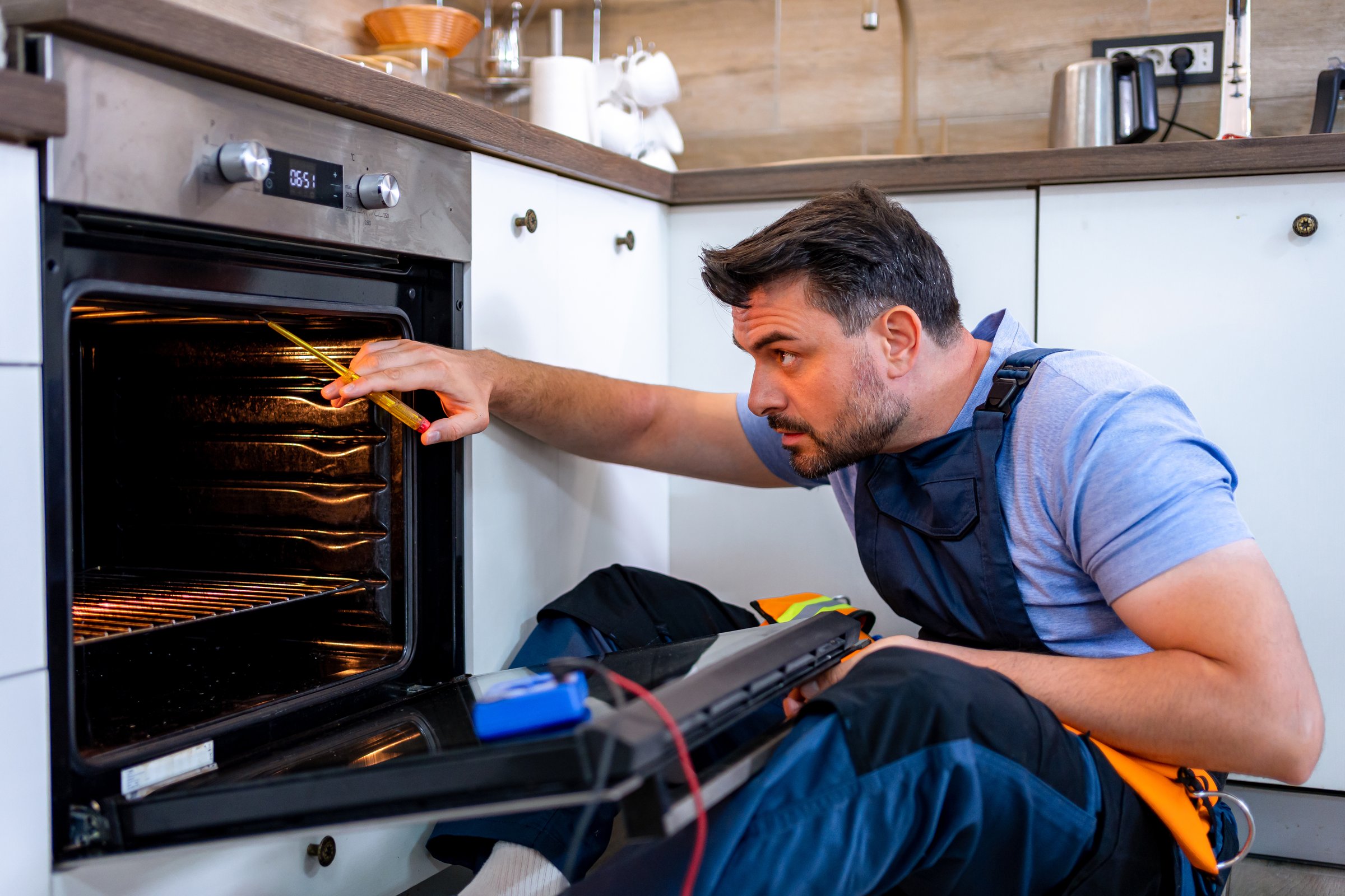 A technician inspects and repairs an oven in a contemporary kitchen during daytime.