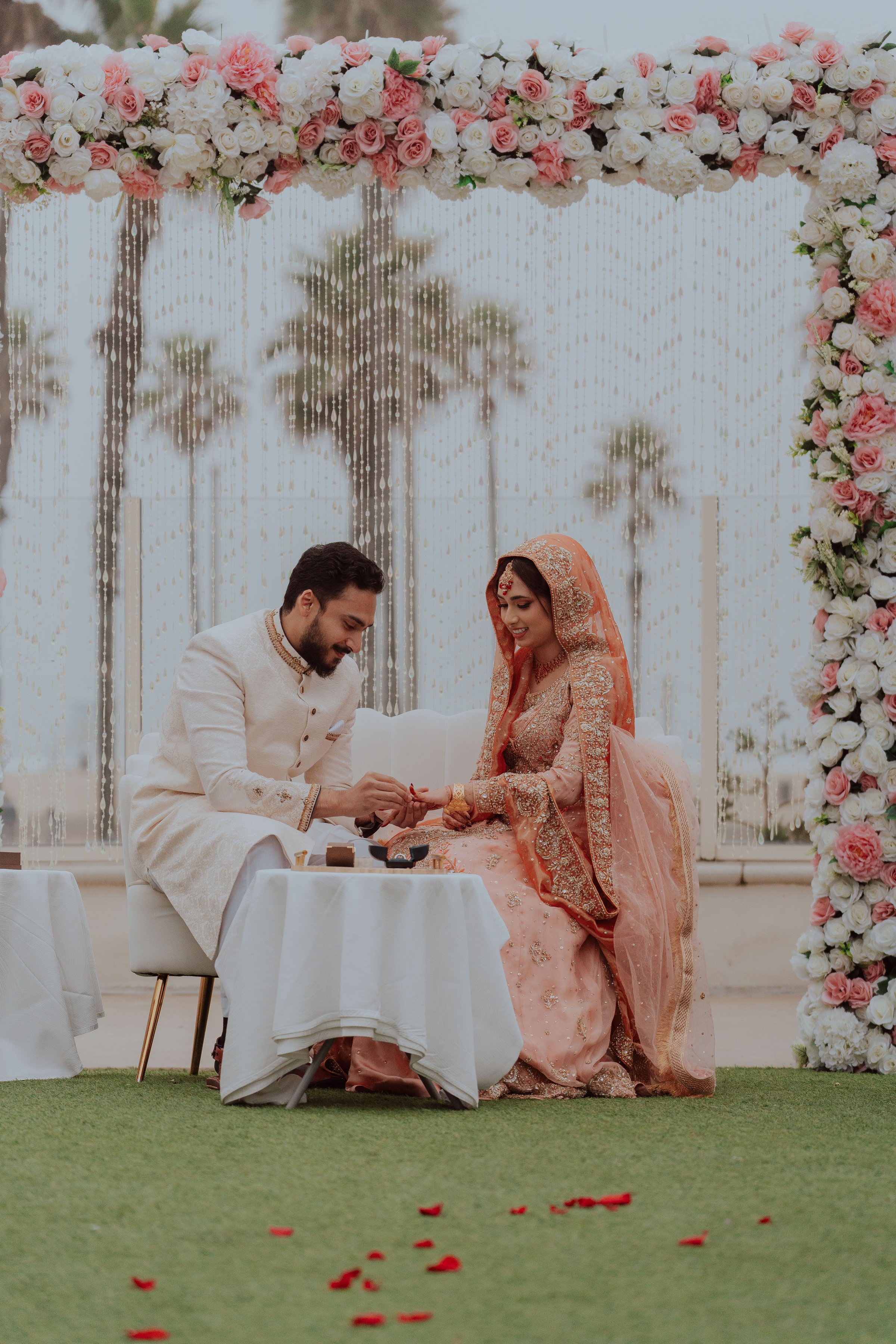 Bride and groom in traditional attire during a wedding ceremony, surrounded by a floral backdrop and palm trees.