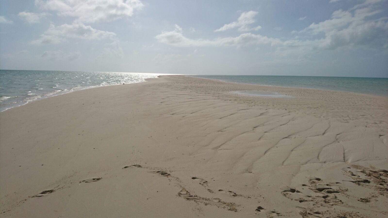 Deserted sandy beach extending into the ocean under a partly cloudy sky, with footprints and a small water puddle visible.