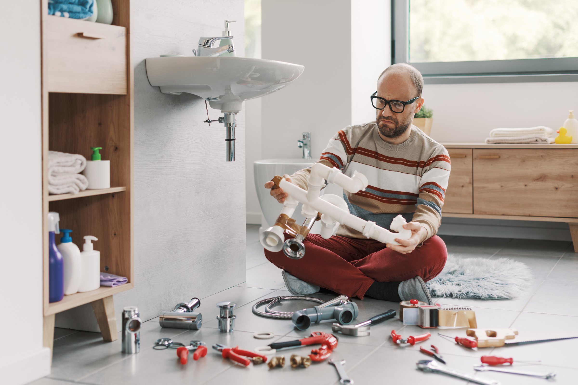 Frustrated man repairing his bathroom sink at home, he is holding plumbing pipes and feeling confused