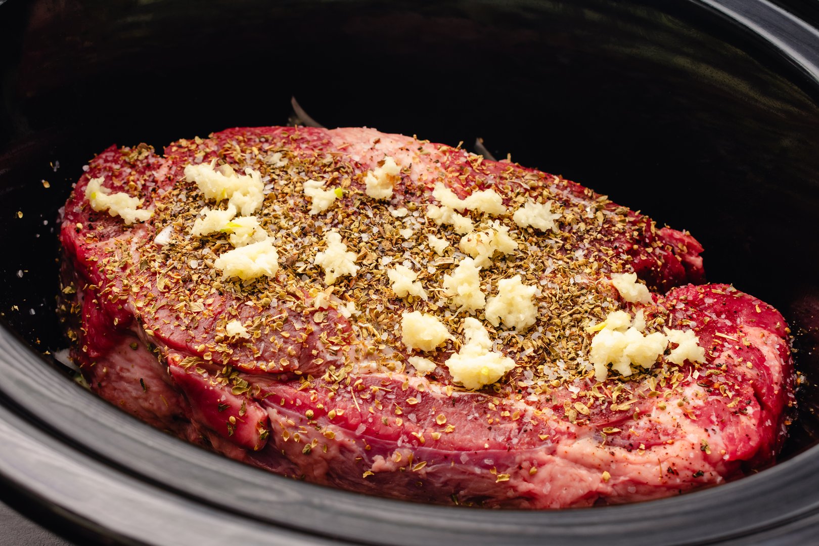 Uncooked beef chuck roast in a crockpot with dried oregano, minced garlic, salt, and pepper