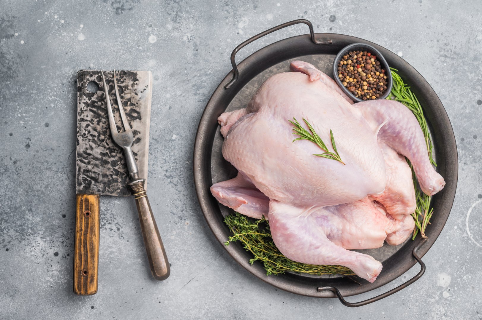 Raw whole chicken on a rustic metal tray with fresh rosemary, thyme, and peppercorns on a gray concrete background, ready for cooking