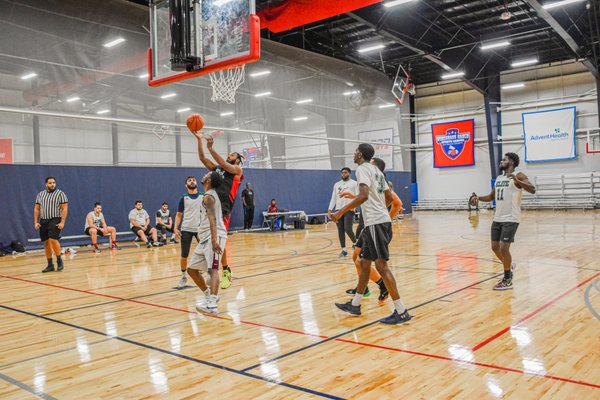 Indoor basketball game with players in mid-action near the hoop, one attempting a layup, referees and bench visible.