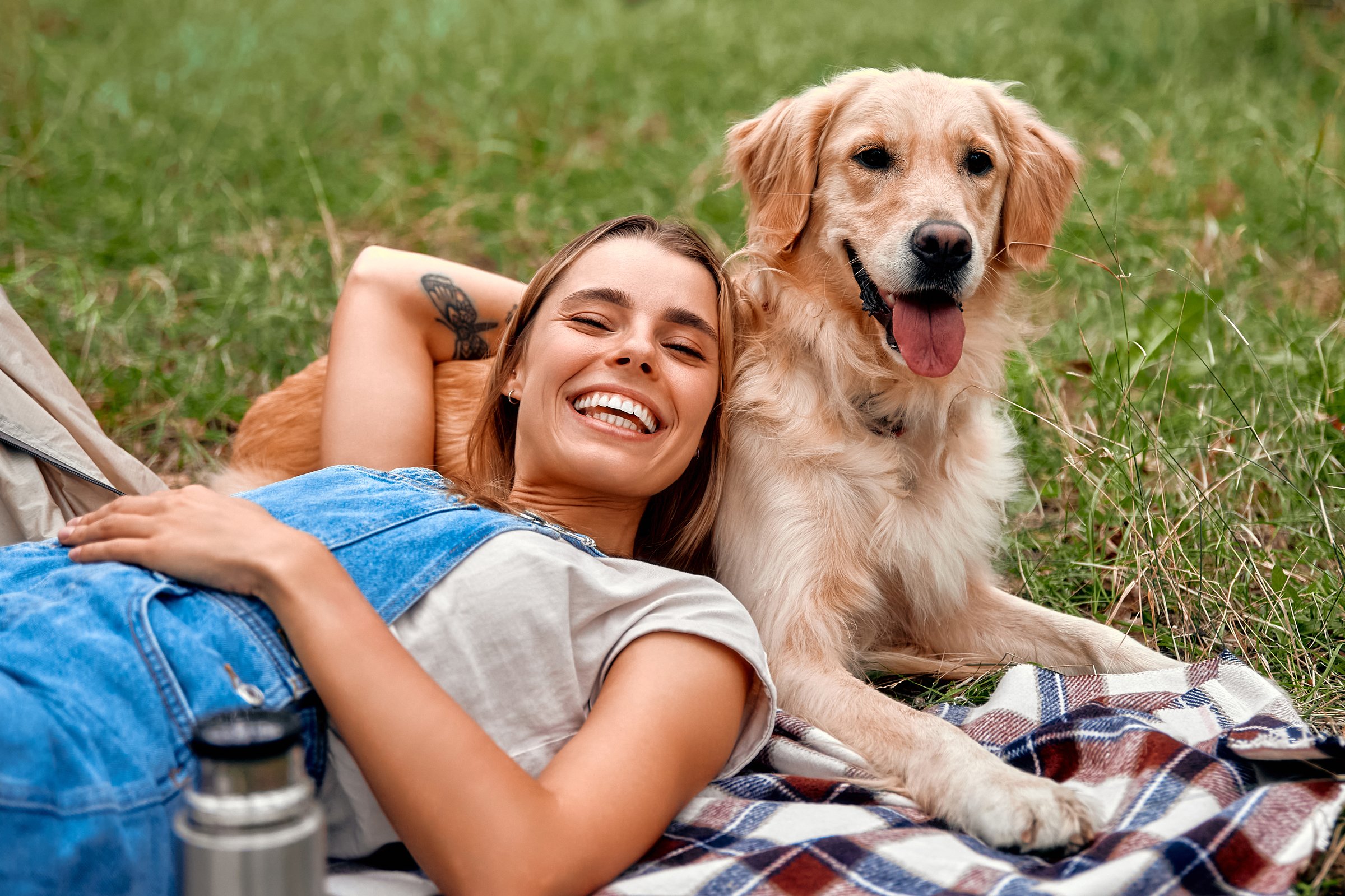 A cheerful woman is fully immersed in enjoying a beautiful sunny day while lying comfortably on the lush grass with her playful golden retriever, capturing blissful, joyful moments in nature