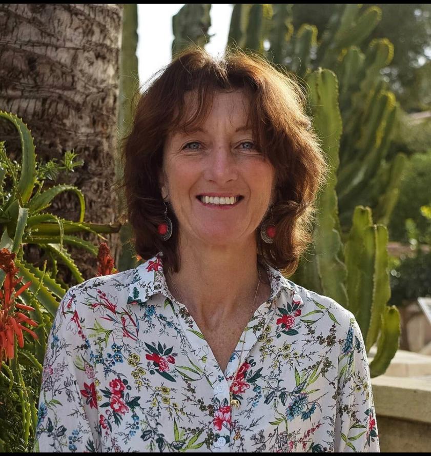 Woman with short brown hair in a floral shirt smiling in front of plants, including cacti and trees, on a sunny day.