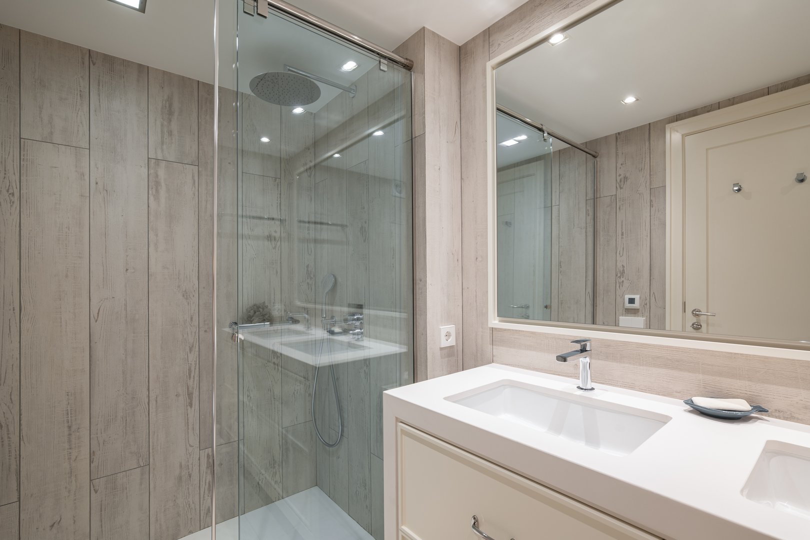 Spacious bathroom with bright artificial lighting, large mirror reflecting front door and pair of snow-white sinks built into light beige cabinet. Shower enclosure with blue glass splash guard