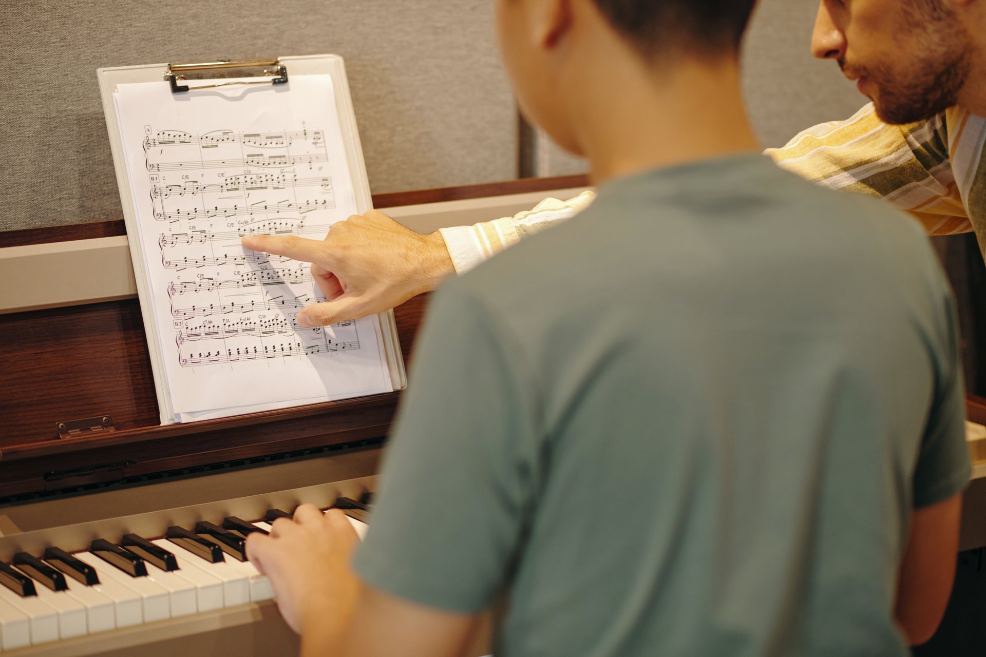 Close-up of a piano teacher guiding student using musical sheet in music classroom, both focusing on the notes. Student holding sheet while teacher points at music notes