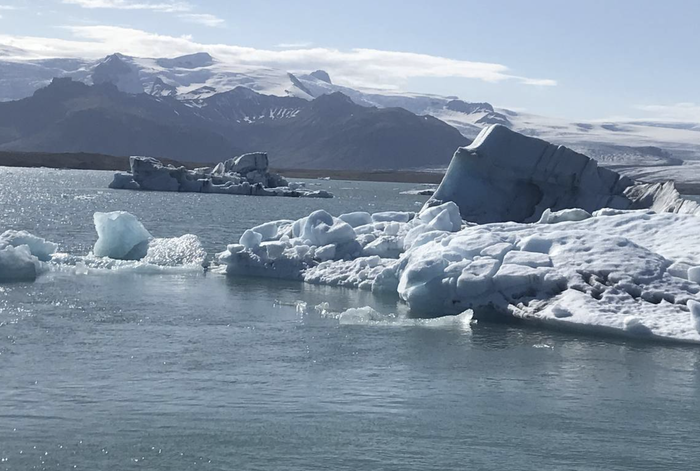 Icy landscape with large icebergs floating in a tranquil body of water, surrounded by snow-capped mountains under a clear sky.