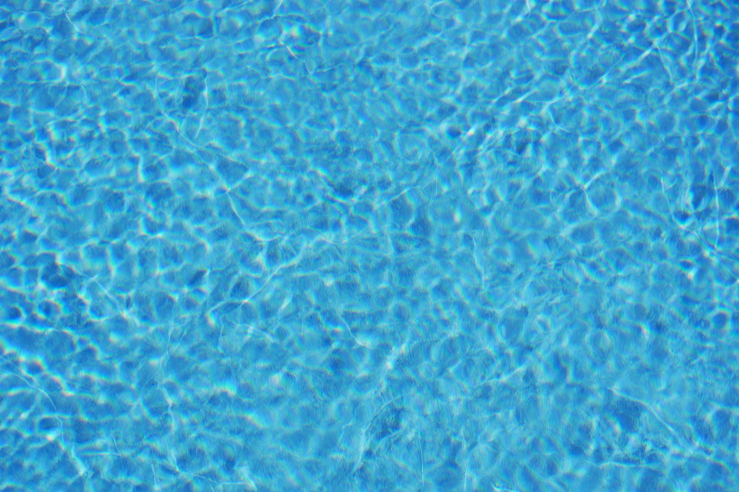 Vibrant close-up of a crystal clear blue swimming pool surface with bright sunlight reflections creating ripple patterns. Background for summer, vacation, leisure or aquatic-themed designs