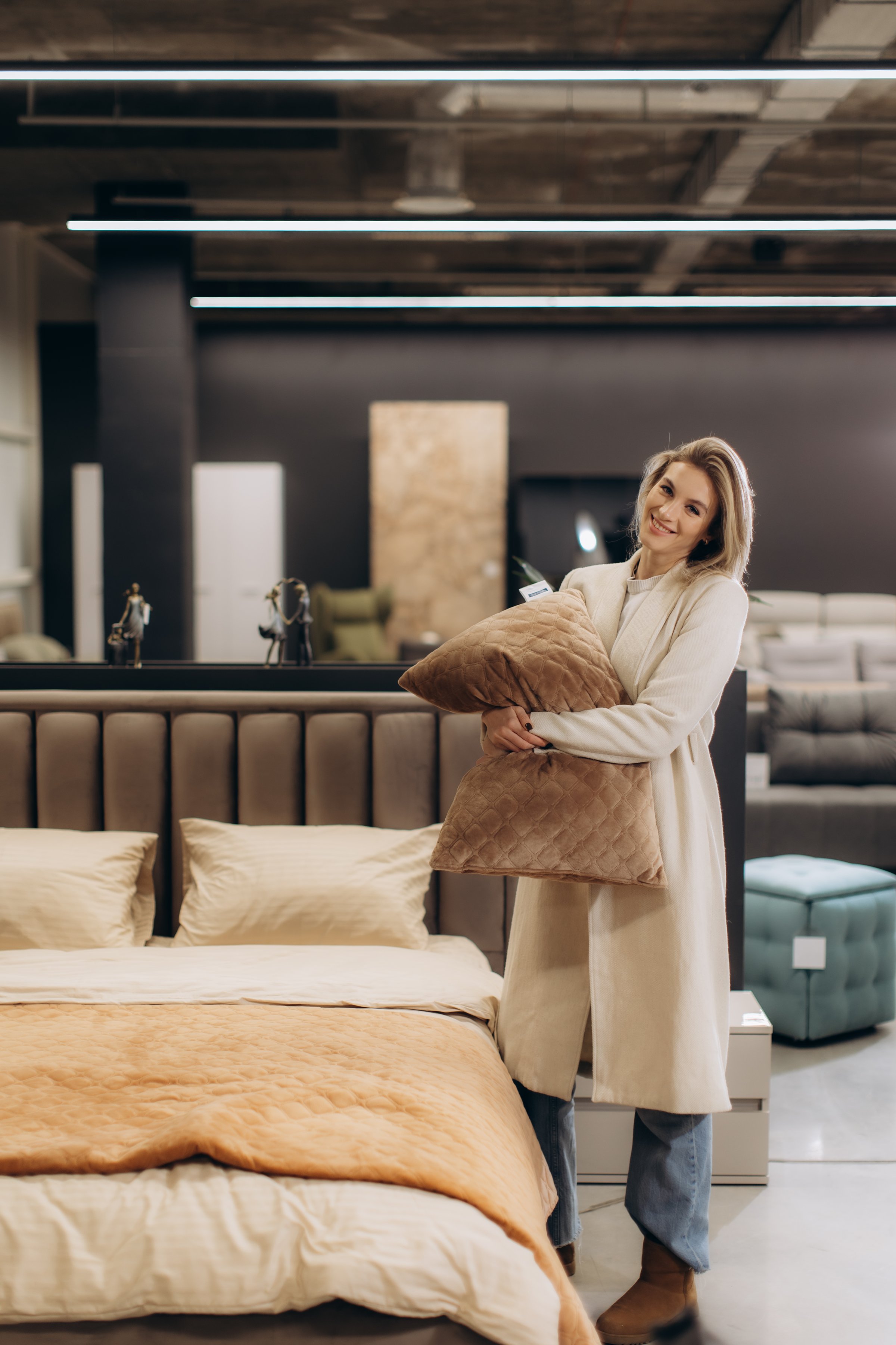 A cheerful woman poses with a pillow near a stylish bed in a furniture store, suggesting comfort and contemporary interior design. The setting highlights luxury home decor and the shopping experience.