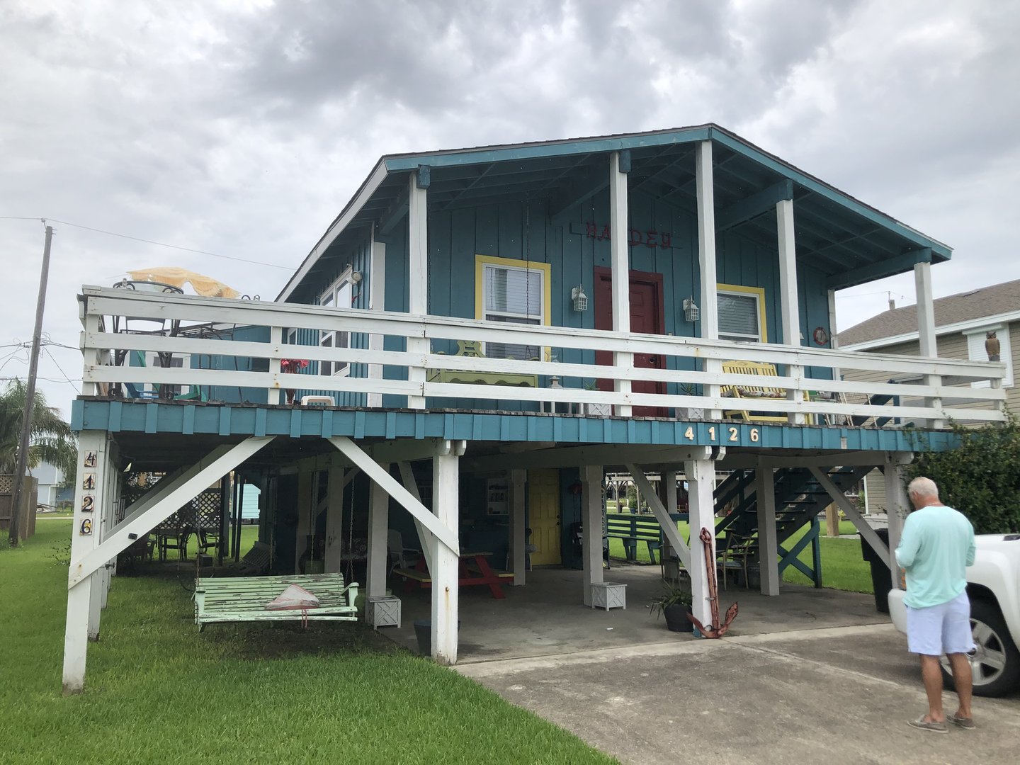Elevated blue wooden house with a porch and a white railing. A person stands on the driveway below. Overcast sky above.