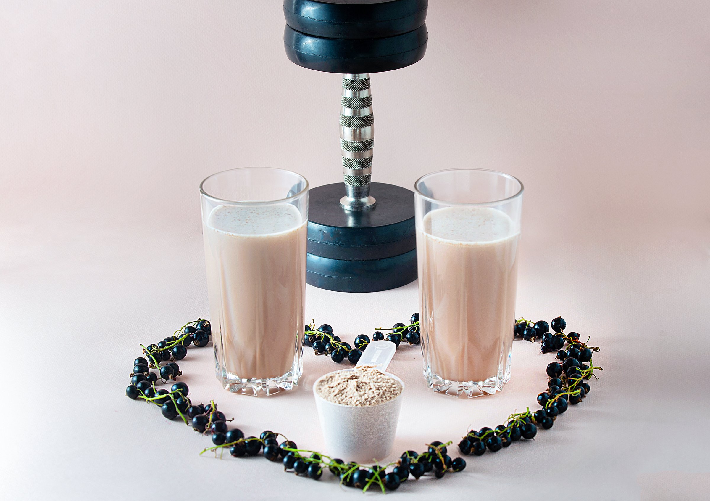 Against a light background, two glass glasses with a ready-made protein drink, a heart of black currant lined around them, in the background of a dumbbell.