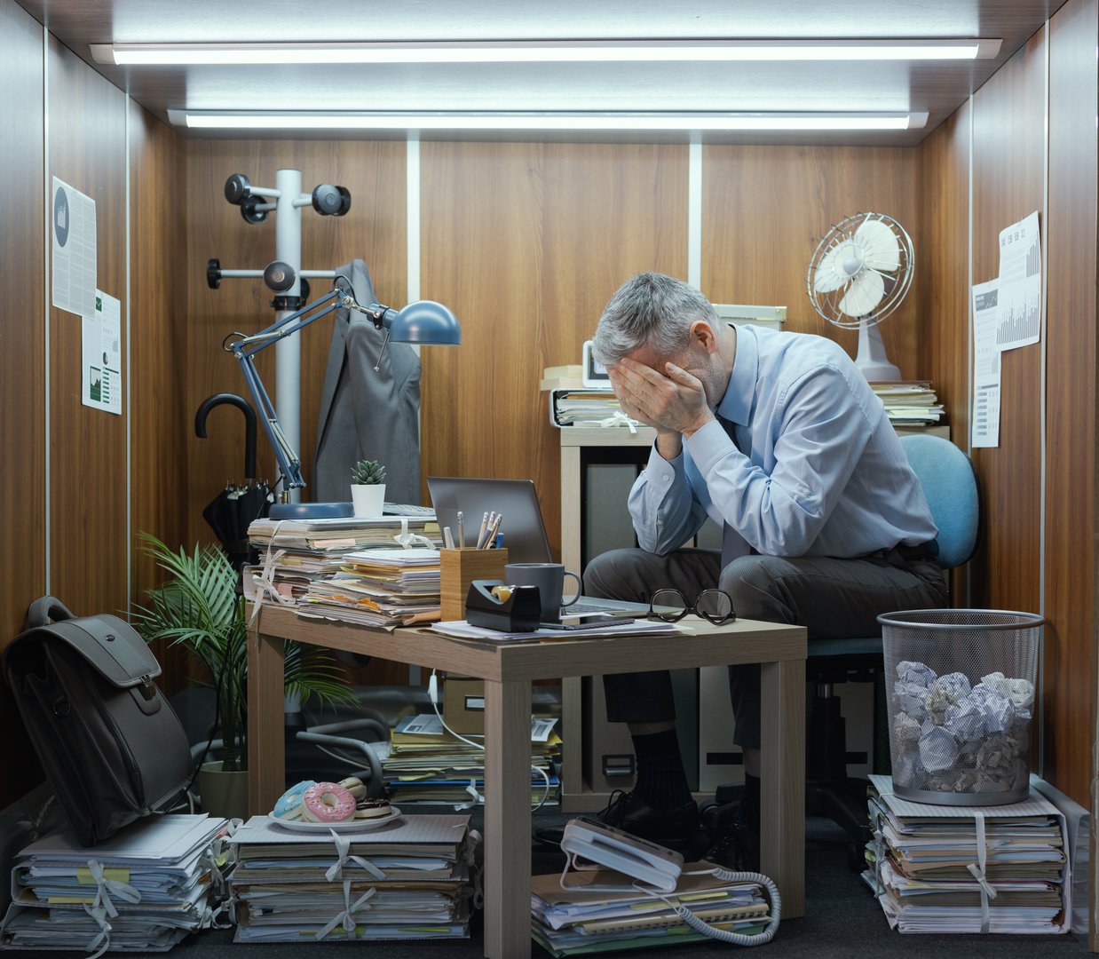 Frustrated company employee working in a small messy office, he is sitting at his desk and crying with head in hands