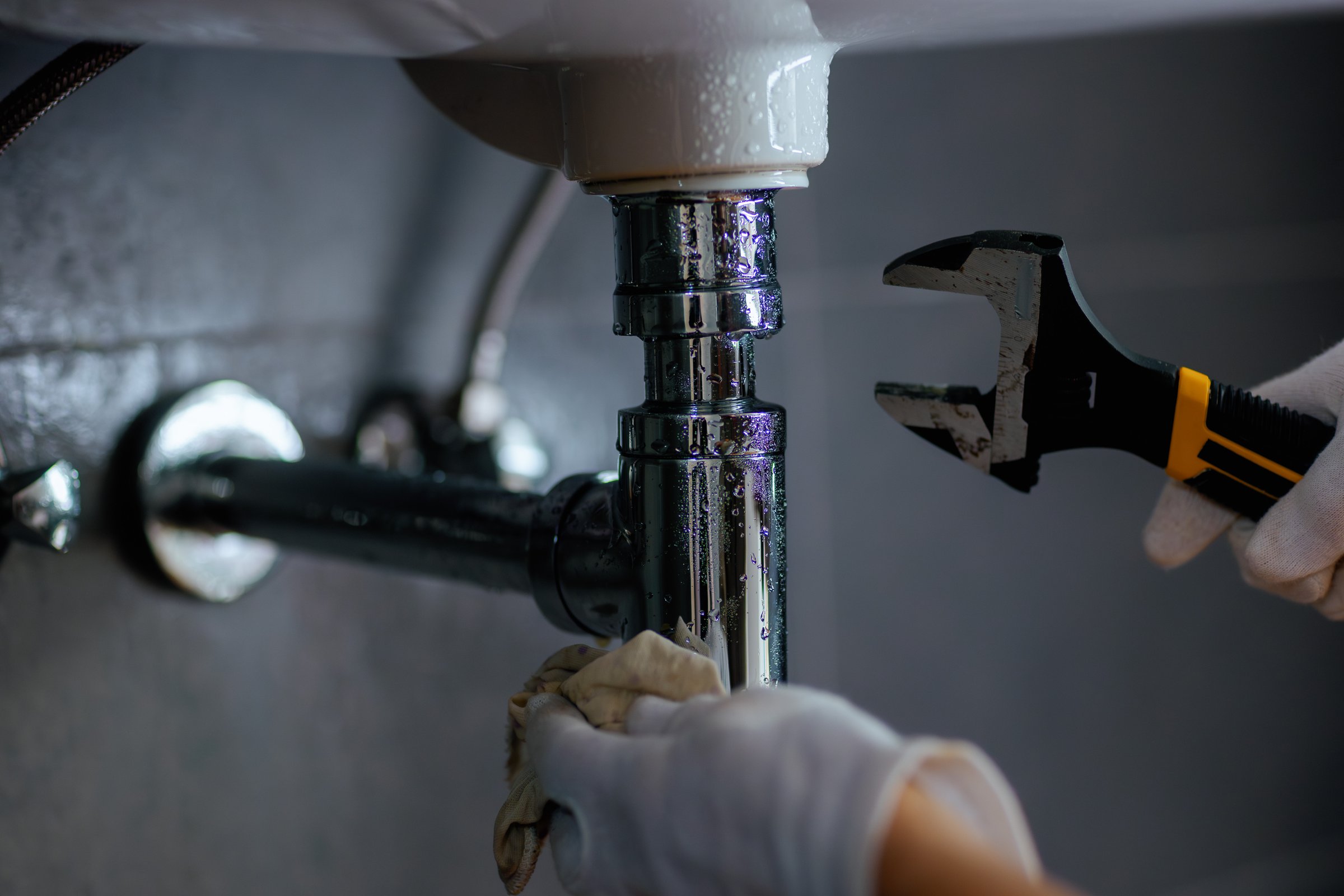 A person in white gloves uses a wrench and cloth to work on chrome plumbing under a sink, suggesting repair and maintenance.