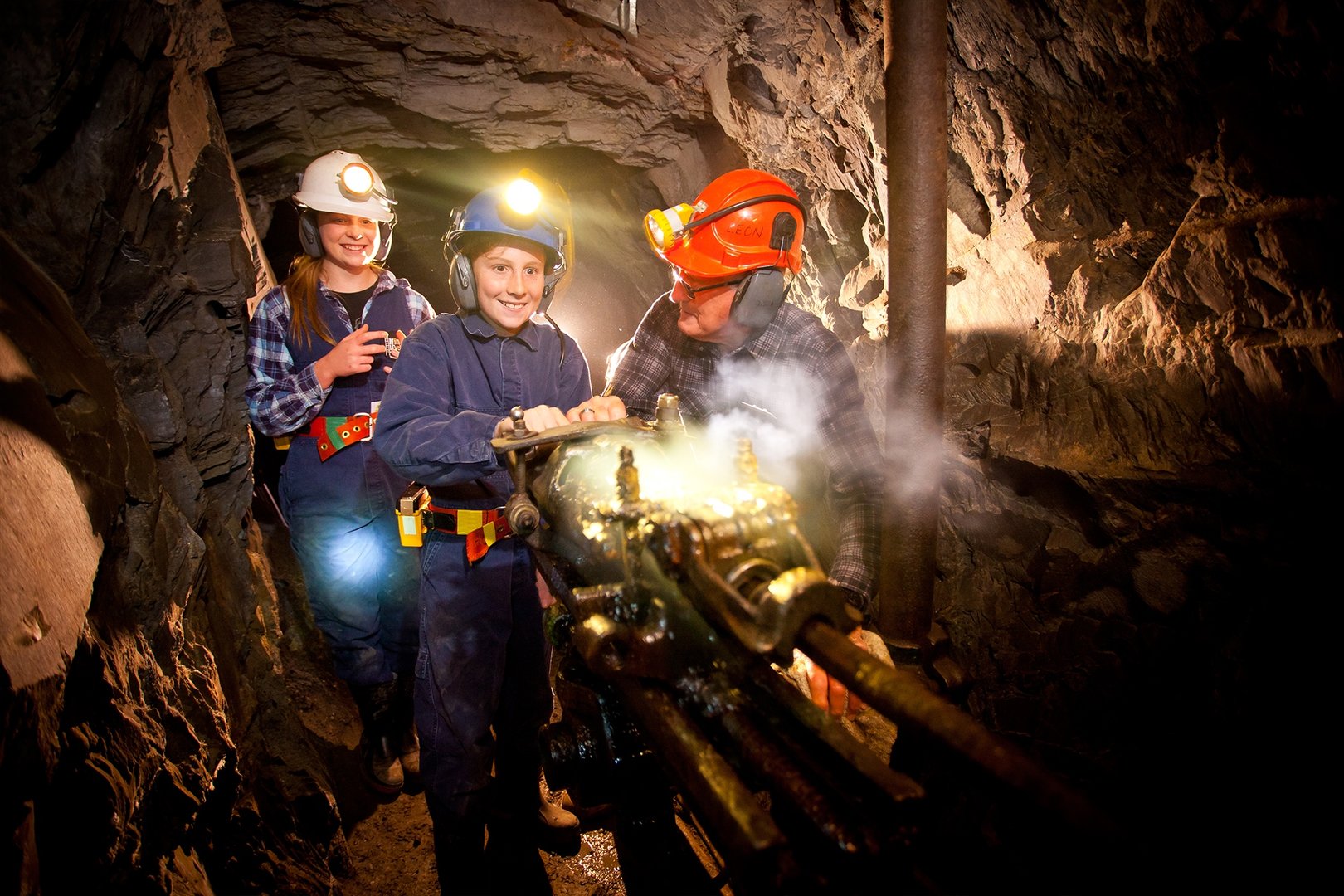 Three people wearing helmets operating a mining drill inside a dimly lit underground tunnel.