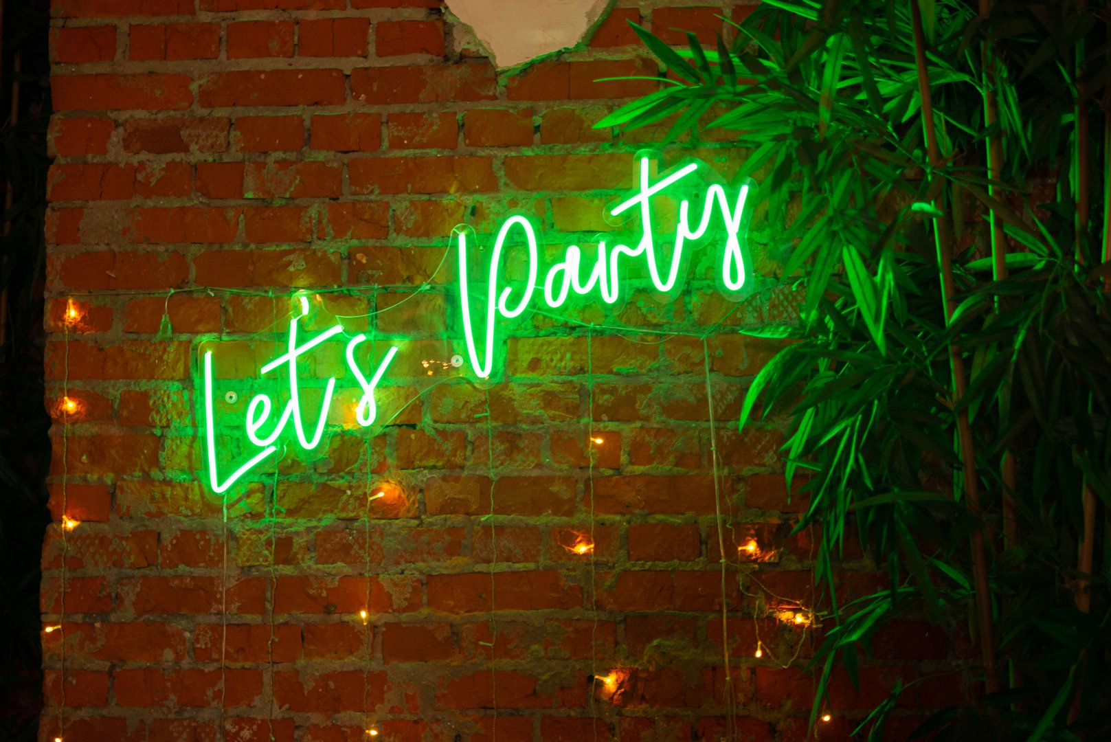 A vibrant green neon sign reads let's party, set against a rustic brick wall adorned with string lights and lush green leaves, creating a festive atmosphere.
