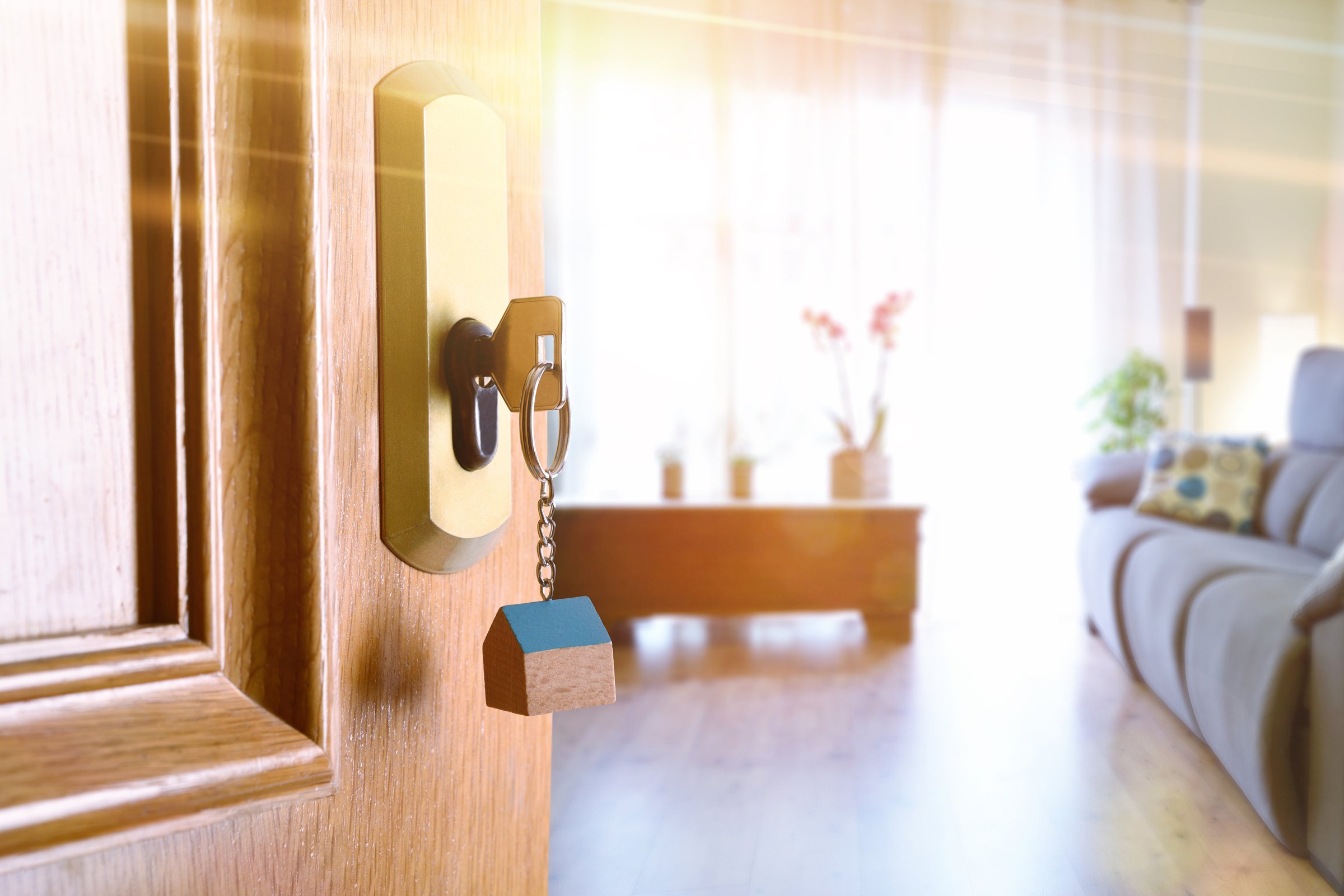 Open entrance door detail of a house with keys in the lock and furnished living room in the background with golden light effect.