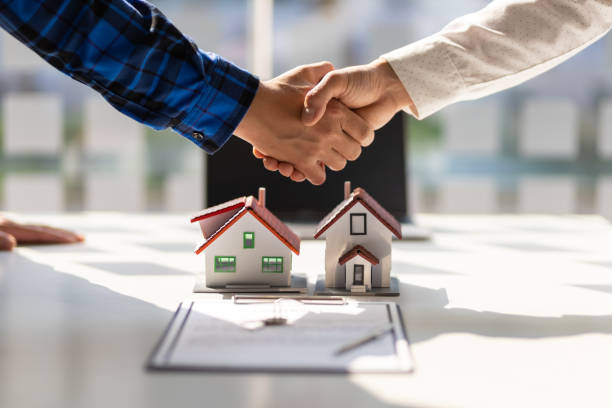 Two people shaking hands over miniature house models on a desk, symbolizing real estate agreement or transaction.