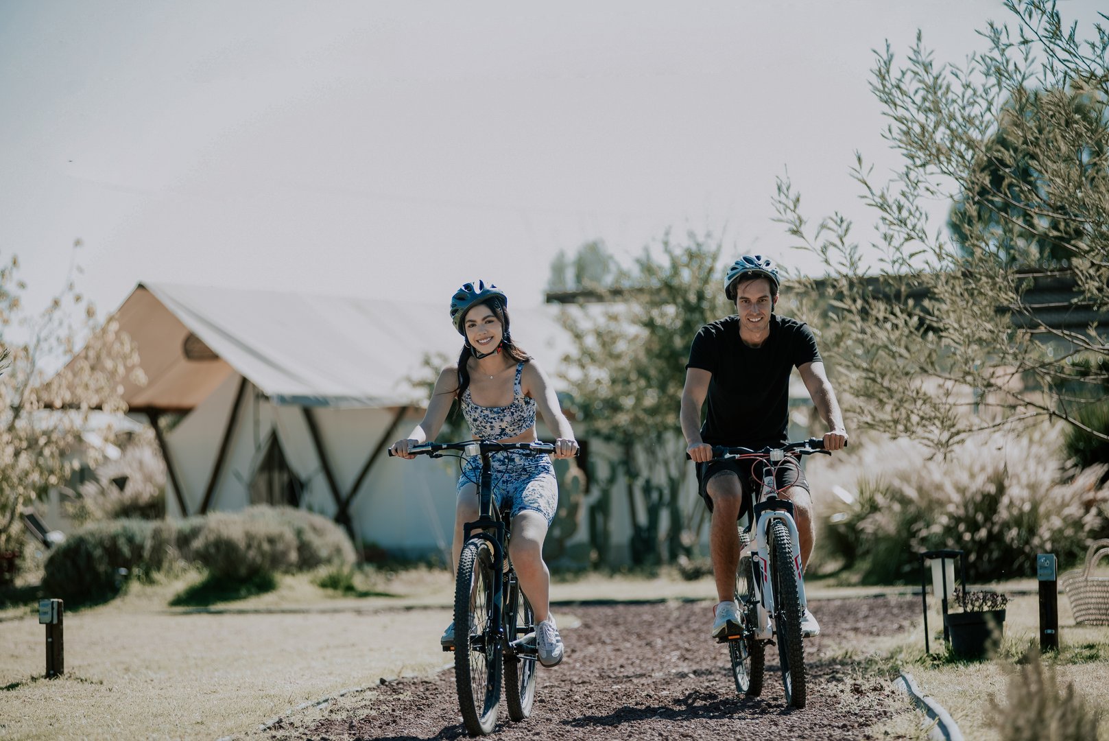 Two people wearing helmets riding bicycles on a gravel path next to tents in a sunny outdoor campsite.