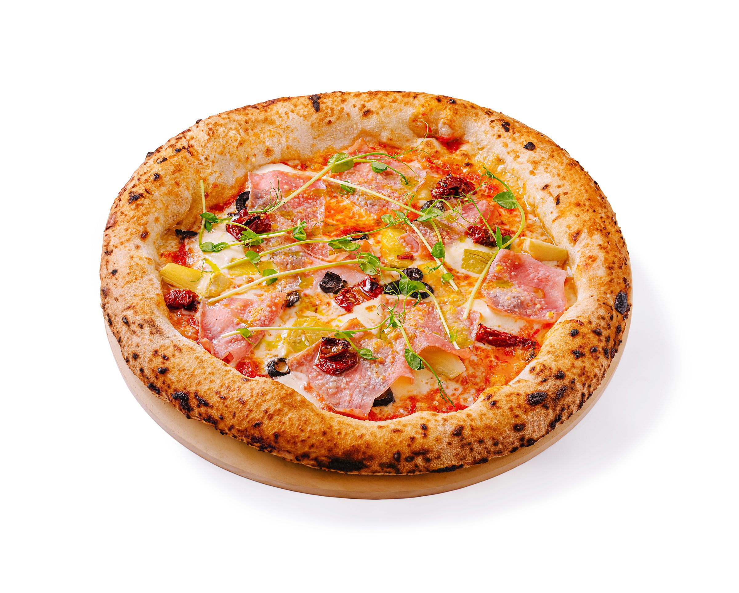 A freshly baked wood-fired pizza topped with ham, olives, and herbs is presented on a wooden platter. The golden crust is perfectly charred, showcasing a blend of ingredients.