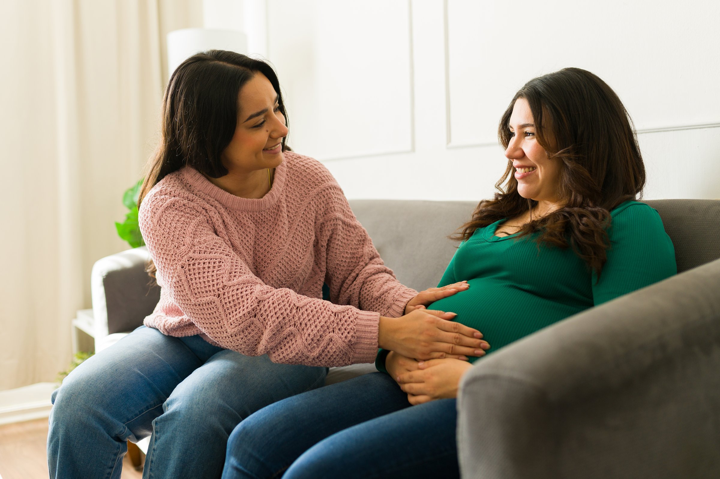 Pregnant woman gently touching belly, feeling fetal movement while conversing with supportive doula during prenatal consultation