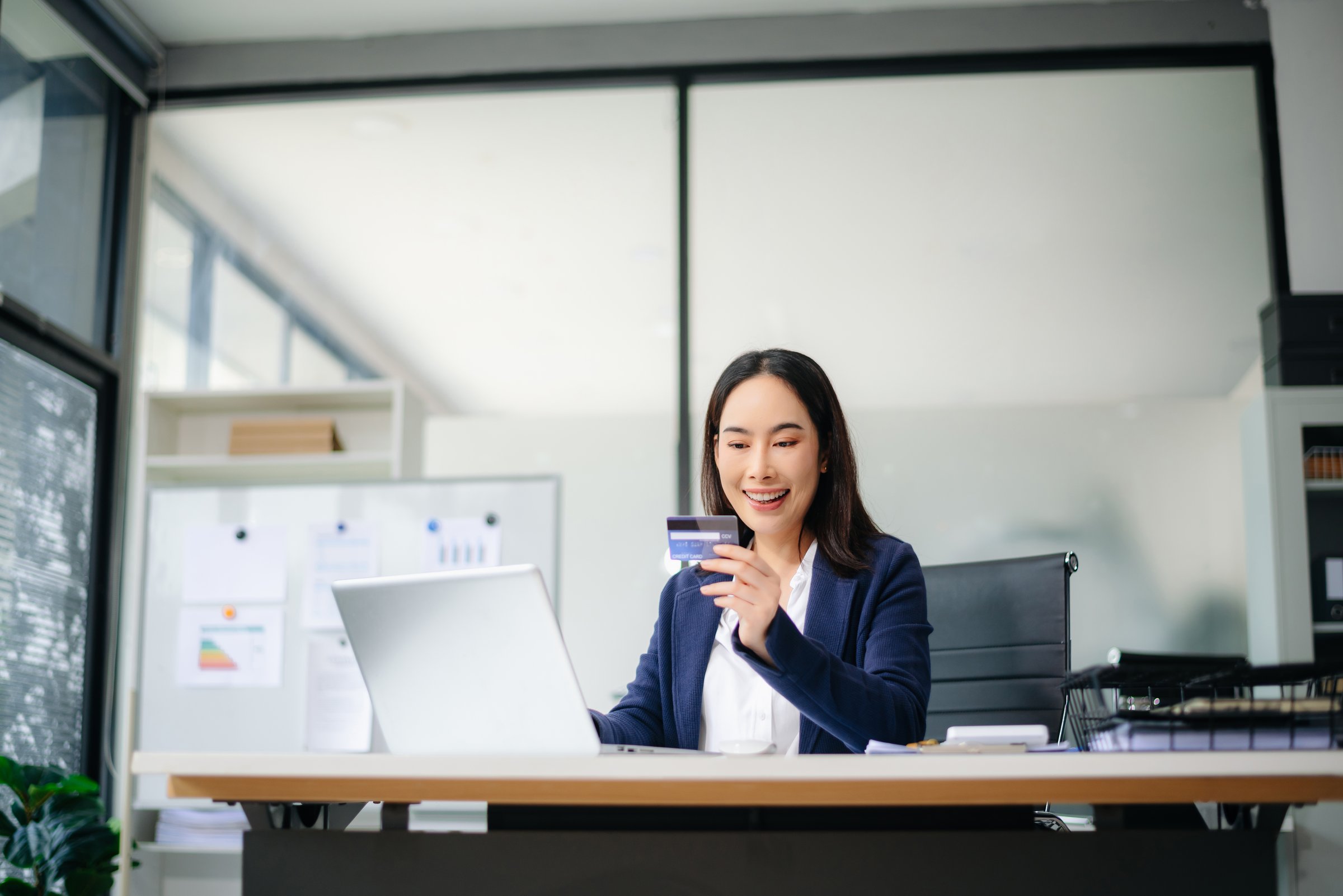 Confident Asian businesswoman smiling while using smartphone and credit card at office desk.