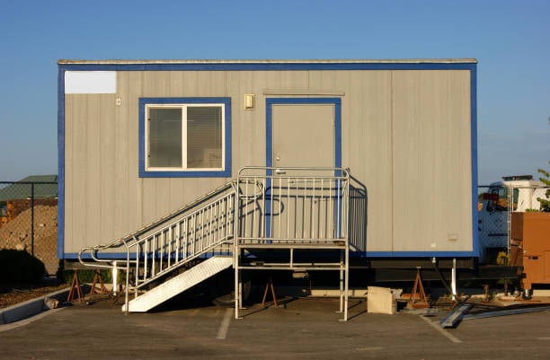 A portable office trailer with stairs and a small window, set up in an outdoor area with a clear blue sky backdrop.