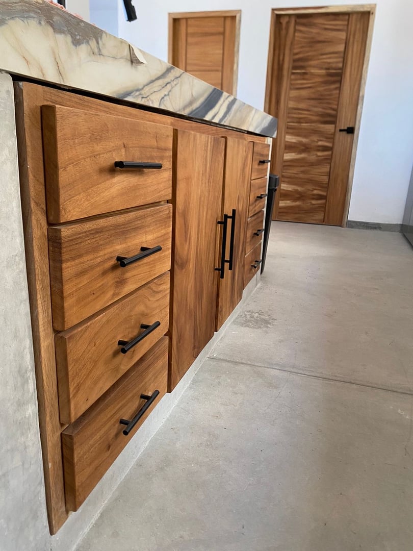 Wooden kitchen cabinets with black handles and a marble countertop on a concrete floor.