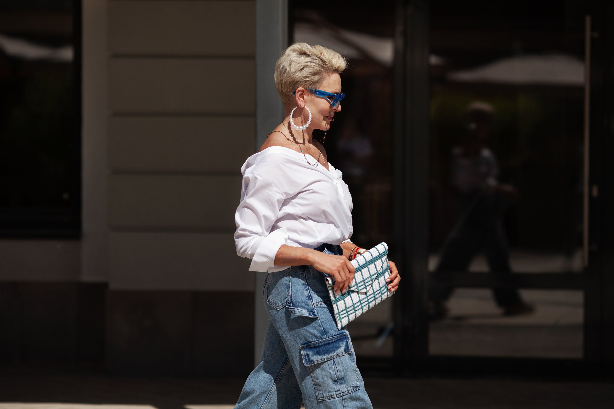 Sophisticated woman with short hair in white shirt and cargo jeans pants, fashionable accessories earrings, stylish glasses and clutch walking outdoors in city. Close up Fashion style.
