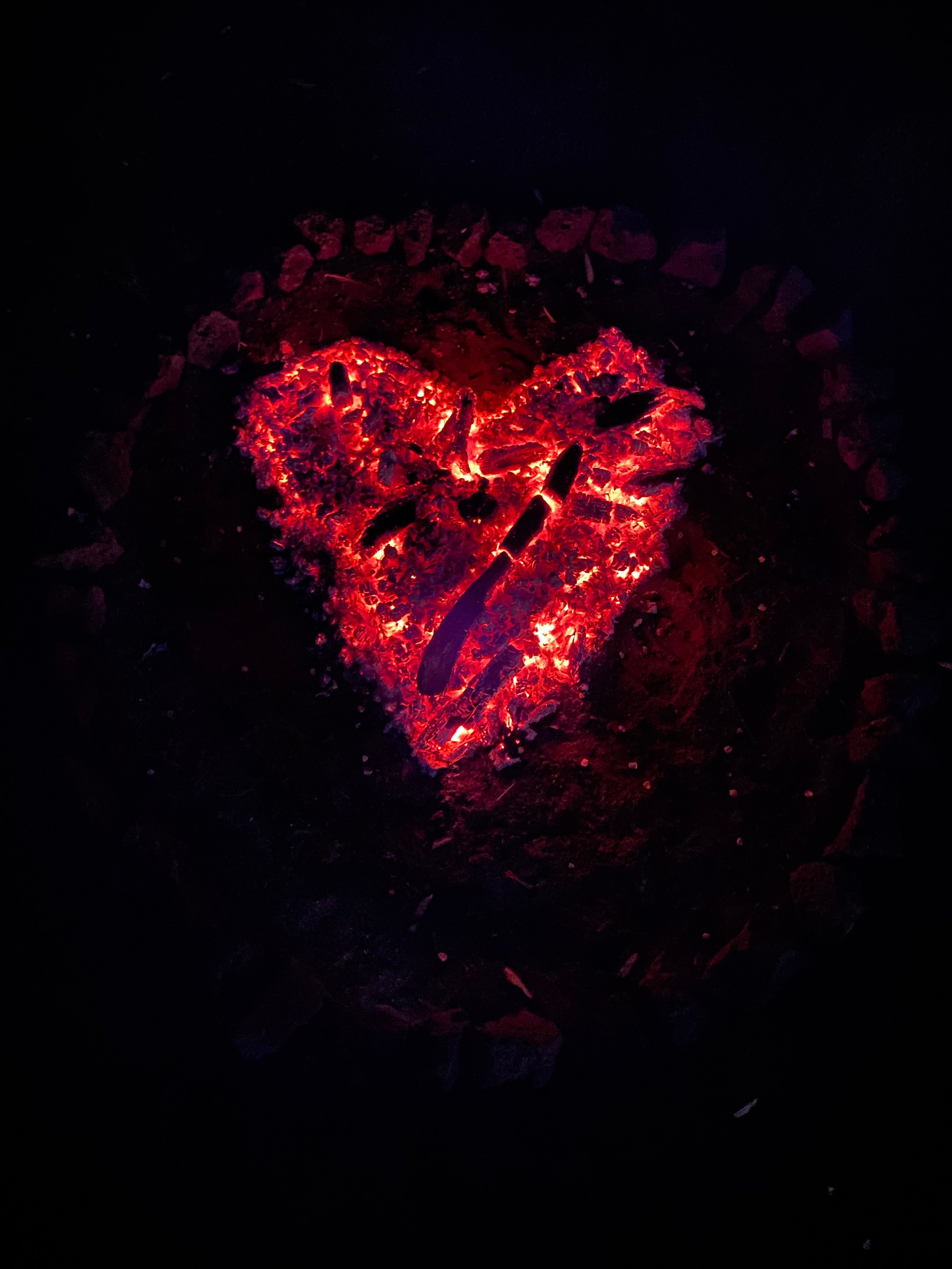 A heart-shaped campfire with glowing embers and ashes, surrounded by darkness, creating a warm and cozy atmosphere.