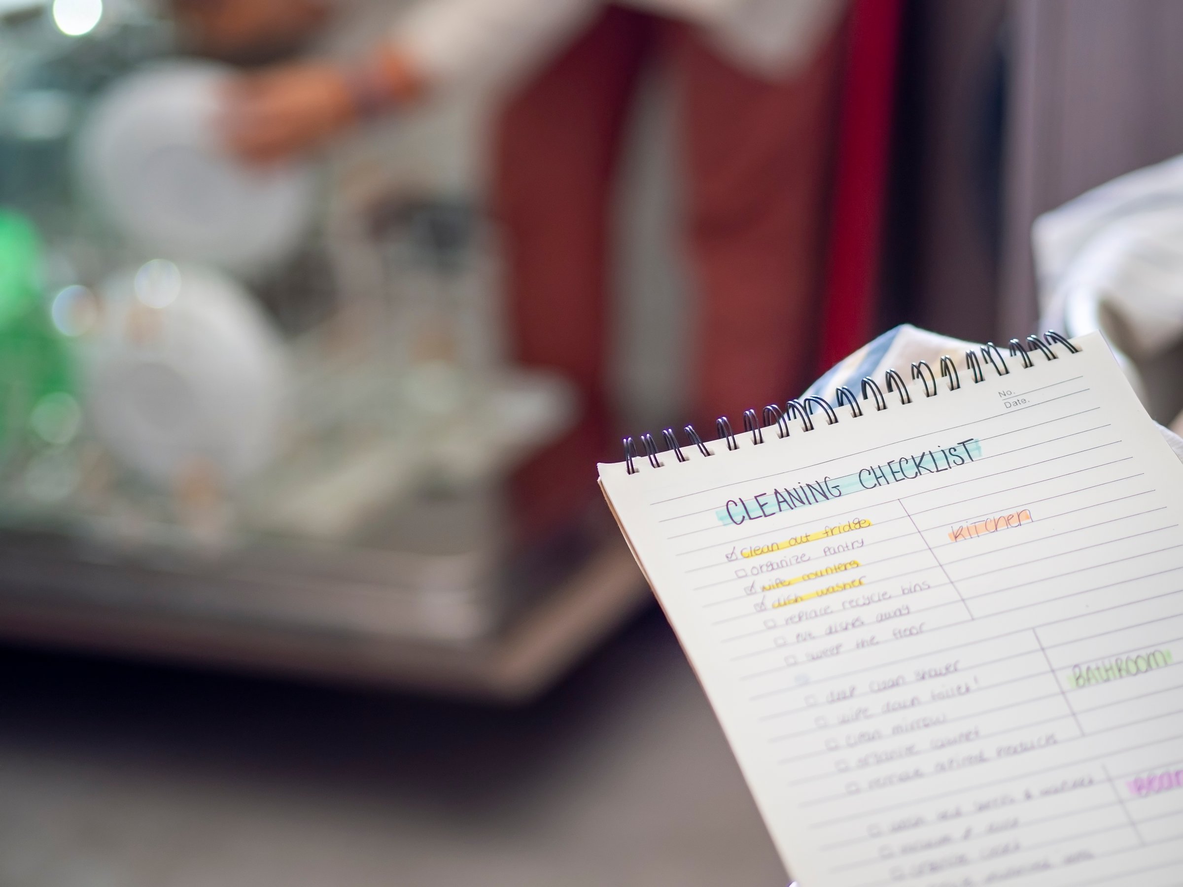 Cleaning checklist for planning routine at home with marks. Woman in the background doing kitchen chores and loading the dishwasher. Organizing home tasks for cleanliness. Domestic life