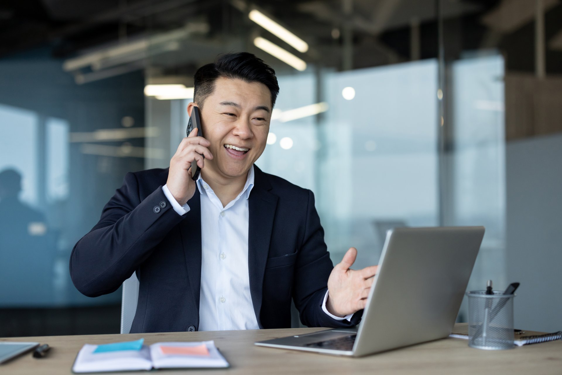 A young Asian man in a suit sits in the office in front of a laptop at a table and talks on a mobile phone with a smile.