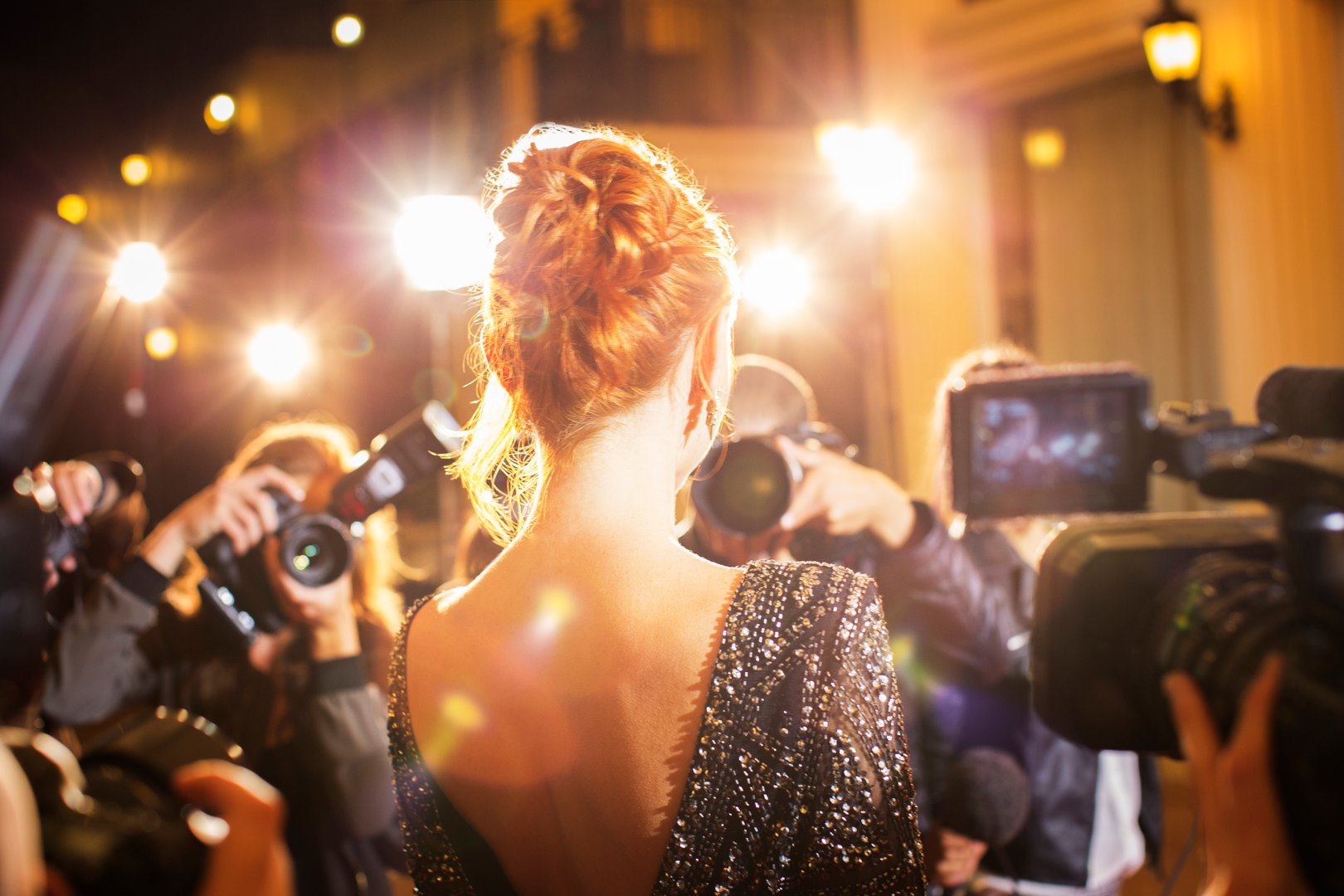 Woman with red hair in sequined dress facing paparazzi photographers with flashing cameras on a red carpet event.