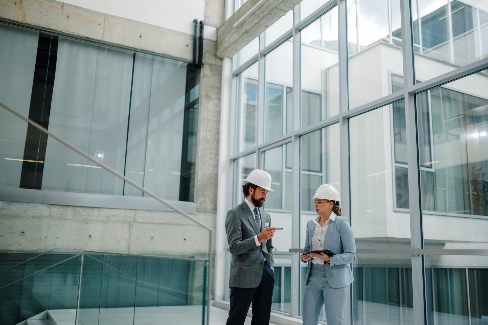 Two architects wearing suits and hardhats are discussing a project in a modern office building