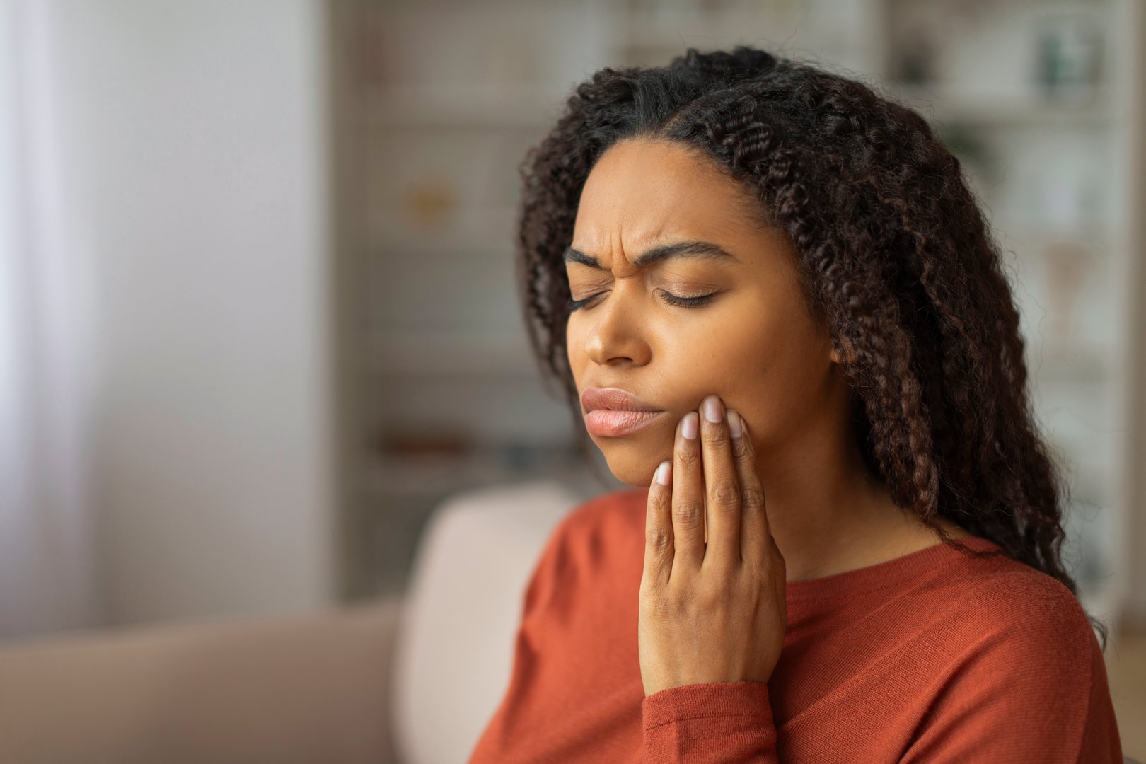 A young black woman pressing her cheek, showing discomfort from a possible toothache, african american female feeling unwell at home, copy space