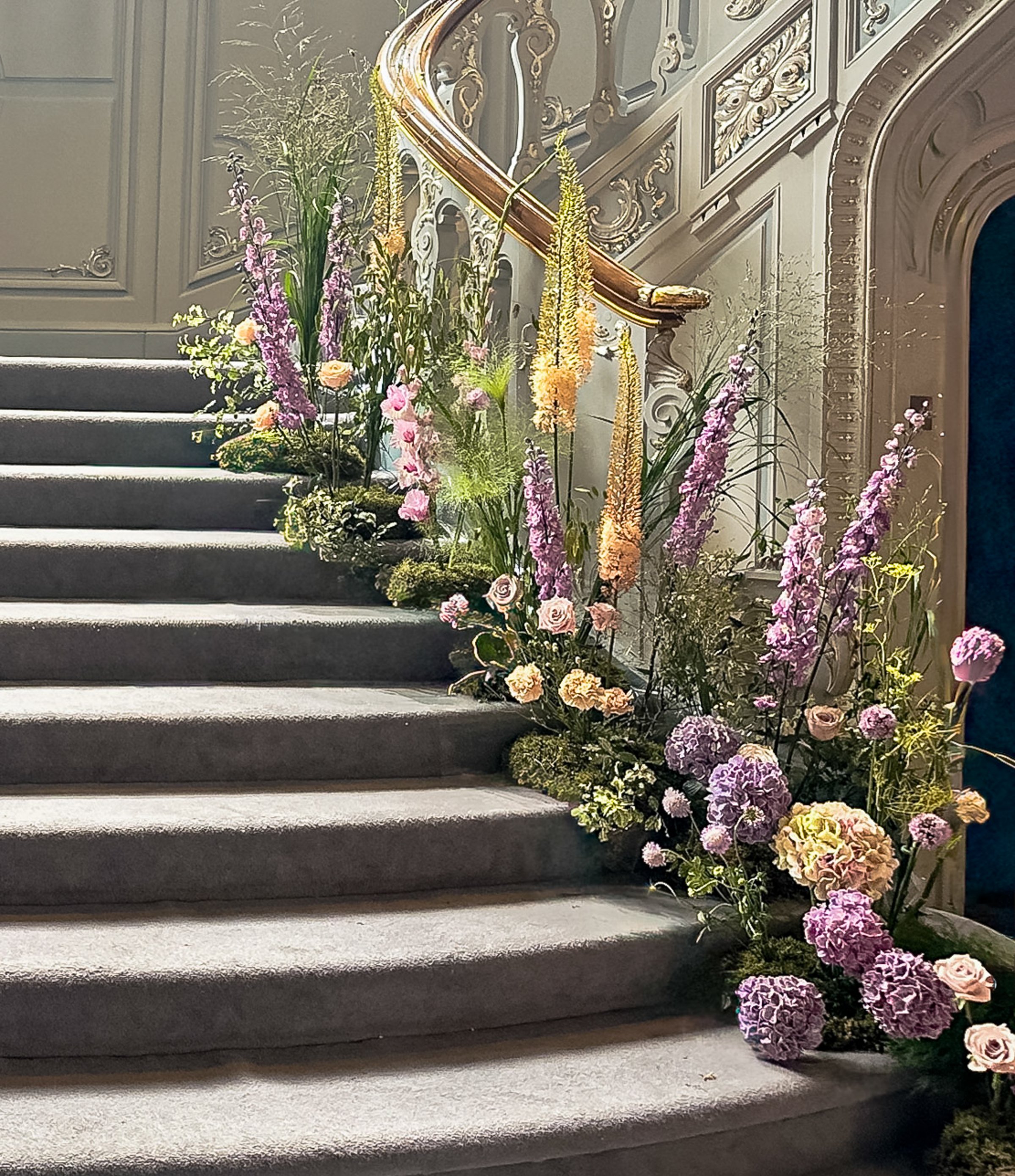A floral installation decorates a classic curved staircase. Fresh roses, hydrangeas, and seasonal flowers are arranged along the steps, creating a luxurious event or wedding decor setting. Vertical image.